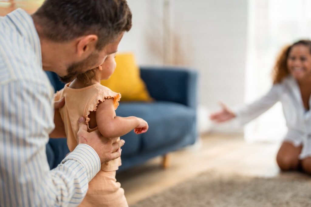 Father helping a toddler take steps toward her smiling mother, showing parents working together in a child custody arrangement.